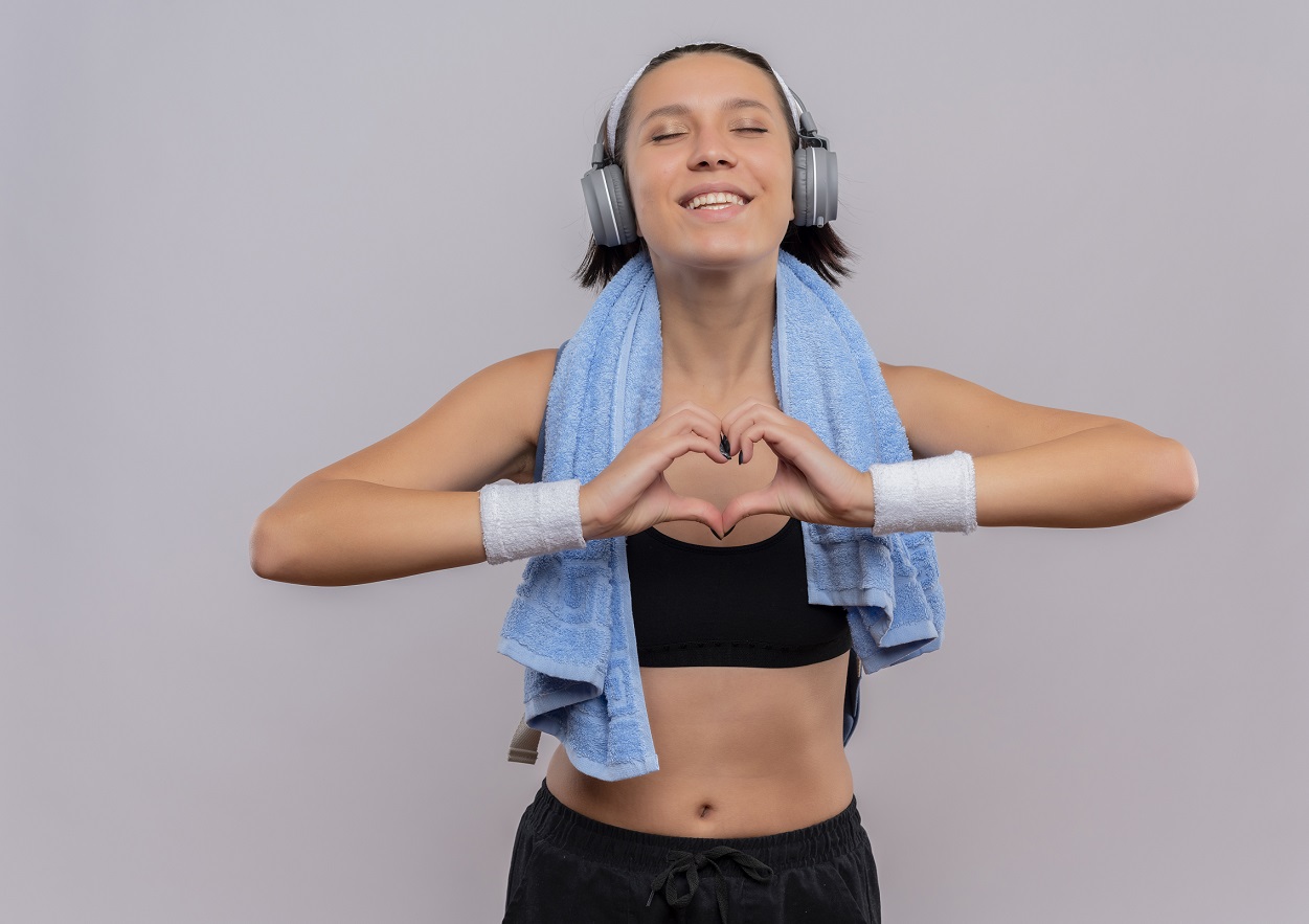 young fitness girl in sportswear with headphones on head and towel on her neck making heart gesture with fingers with closed eyes feeling positive emotions standing over white background